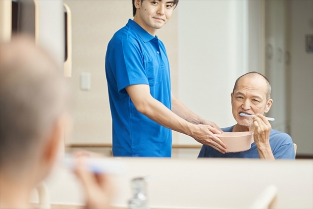 高齢男性が鏡を見ながら歯を磨いている横で、若年男性が洗面器を高齢男性の顎の下で持っている様子