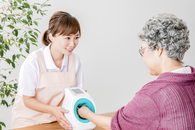 高齢女性が、若年女性が支えている血圧計に左手を入れている様子